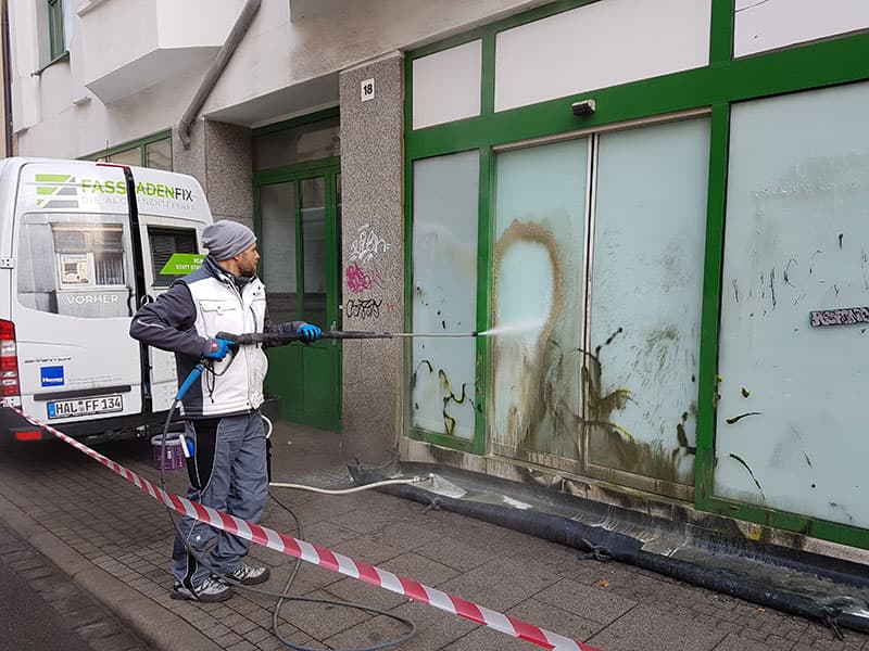FassadenFix-Mitarbeiter entfernt Graffiti von einer Schaufensterfront — mit Auffangsystem am Boden
